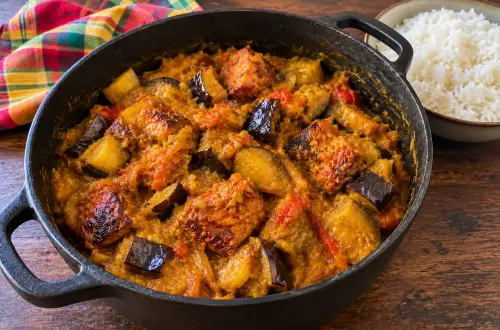 Marmite en fonte de boucané bringelle : morceaux de boucané dorés et cubes d'aubergine avec leur peau, fondus dans une sauce tomate au curcuma, à côté d'un bol de riz blanc