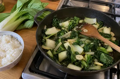 Brèdes chou de Chine sautées à l'ail et à l'oignon dans une sauteuse en fonte sur la gazinière, tiges blanches et feuilles vertes, à côté d'une botte de pak choï et d'un bol de riz