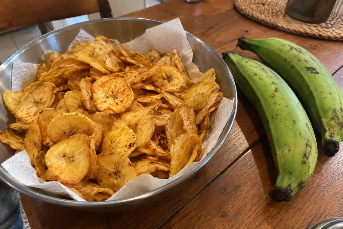 Saladier de chips de banane verte dorées et épicées, à côté de deux bananes plantain vertes