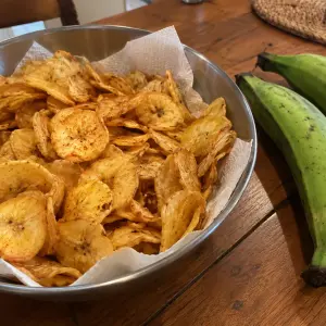 Saladier de chips de banane verte dorées et épicées, à côté de deux bananes plantain vertes