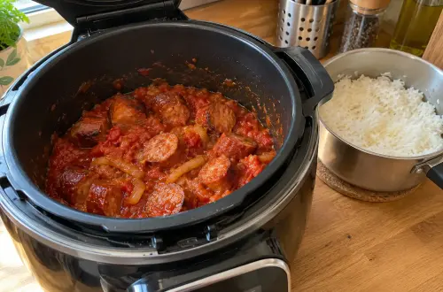 Cookeo ouvert sur un plan de travail en bois, la cuve pleine d'un rougail saucisse en sauce tomate, à côté d'une casserole de riz blanc