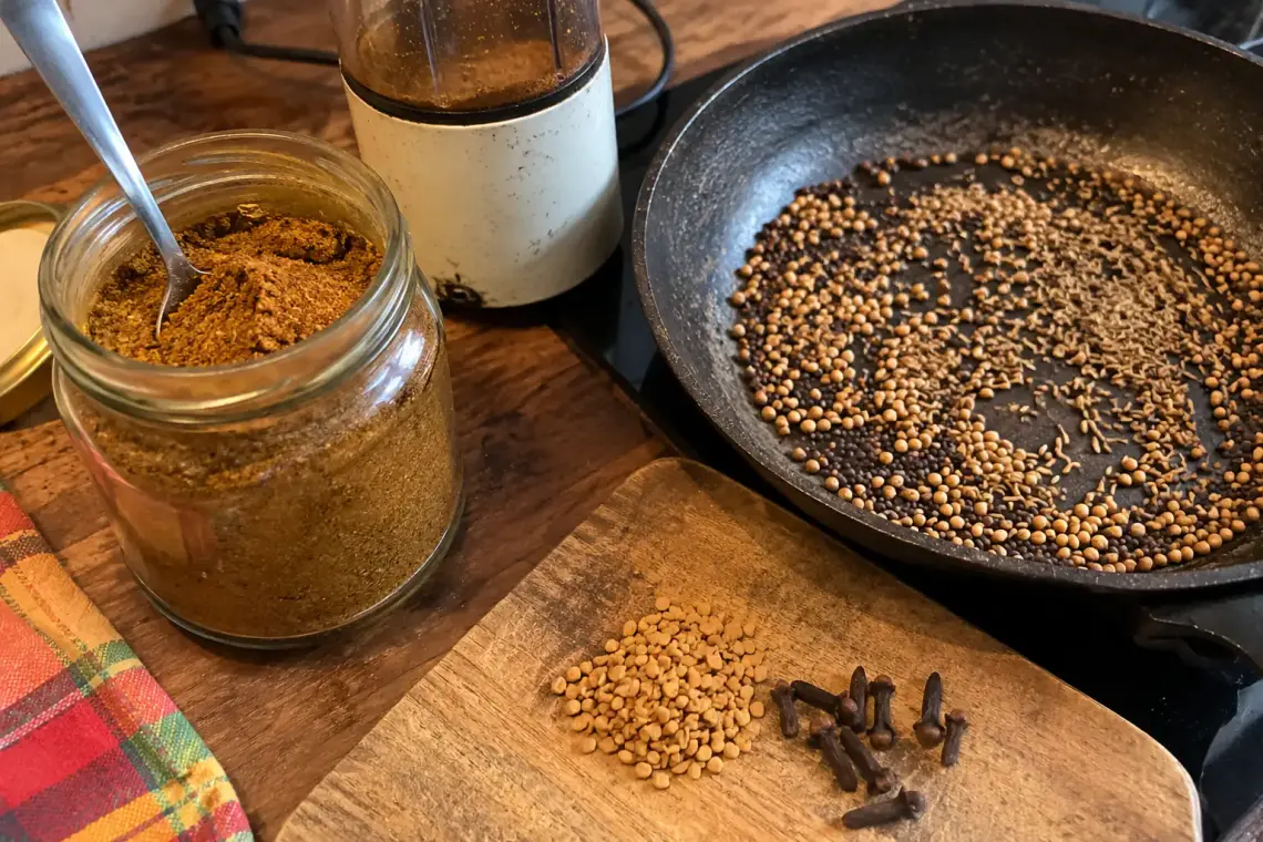 Massalé maison : bocal de poudre brun doré, moulin à café, poêle de graines de coriandre, cumin et moutarde grillées, fenugrec et clous de girofle sur une planche