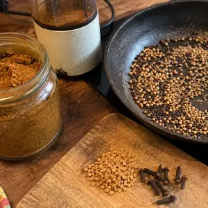 Massalé maison : bocal de poudre brun doré, moulin à café, poêle de graines de coriandre, cumin et moutarde grillées, fenugrec et clous de girofle sur une planche