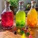 Trois bouteilles de rhum arrangé Dragibus rose, vert et jaune orangé, avec des Dragibus de la même couleur au fond, et des bonbons multicolores sur une table en bois