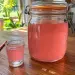 Bocal de rhum arrangé Malabar rose tendre et limpide, avec un petit verre à shot et une paille rayée rose et blanc sur une table en bois