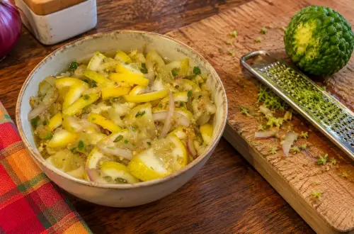 Bol de rougail citron réunionnais, citron jaune coupé avec sa peau, oignon rouge et piment vert, à côté d'un combava à moitié râpé sur une planche