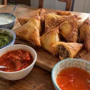 Samoussas au bœuf dorés sur une planche en bois, l'un croqué, entourés de quatre bols de sauces : raïta, chutney vert, sauce tomate pimentée et sauce aigre-douce