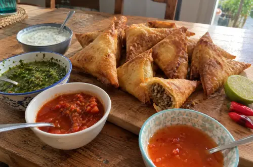 Samoussas au bœuf dorés sur une planche en bois, l'un croqué, entourés de quatre bols de sauces : raïta, chutney vert, sauce tomate pimentée et sauce aigre-douce
