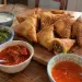Samoussas au bœuf dorés sur une planche en bois, l'un croqué, entourés de quatre bols de sauces : raïta, chutney vert, sauce tomate pimentée et sauce aigre-douce